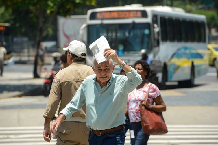 Pessoa idosa se protegendo do calor intenso com um jornal enquanto caminha na rua.