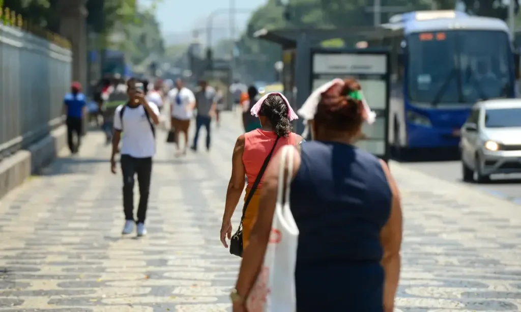 Pessoas caminhando em uma rua durante uma onda de calor, usando roupas leves e chapéus para proteção solar.