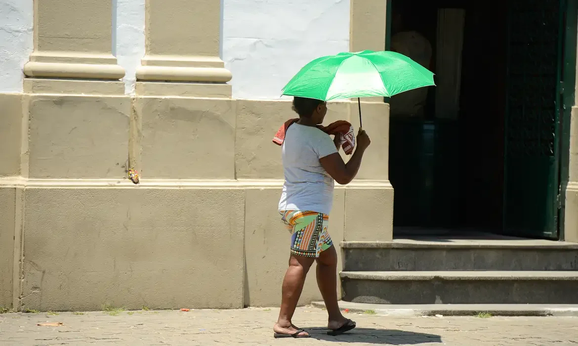 Pessoa caminhando sob forte sol no Nordeste, usando um guarda-sol para proteção contra o calor.