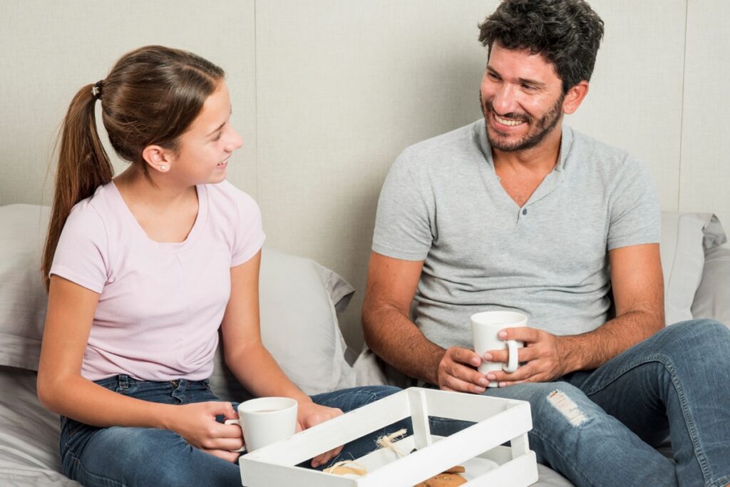 Pai e filha conversando sorridentes enquanto tomam chá, simbolizando a importância do diálogo para o desenvolvimento emocional das crianças.