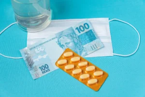 Alerta! Preço dos medicamentos sobe a partir de hoje 1 Dinheiro brasileiro, máscara cirúrgica e cápsulas de remédio sobre fundo azul.