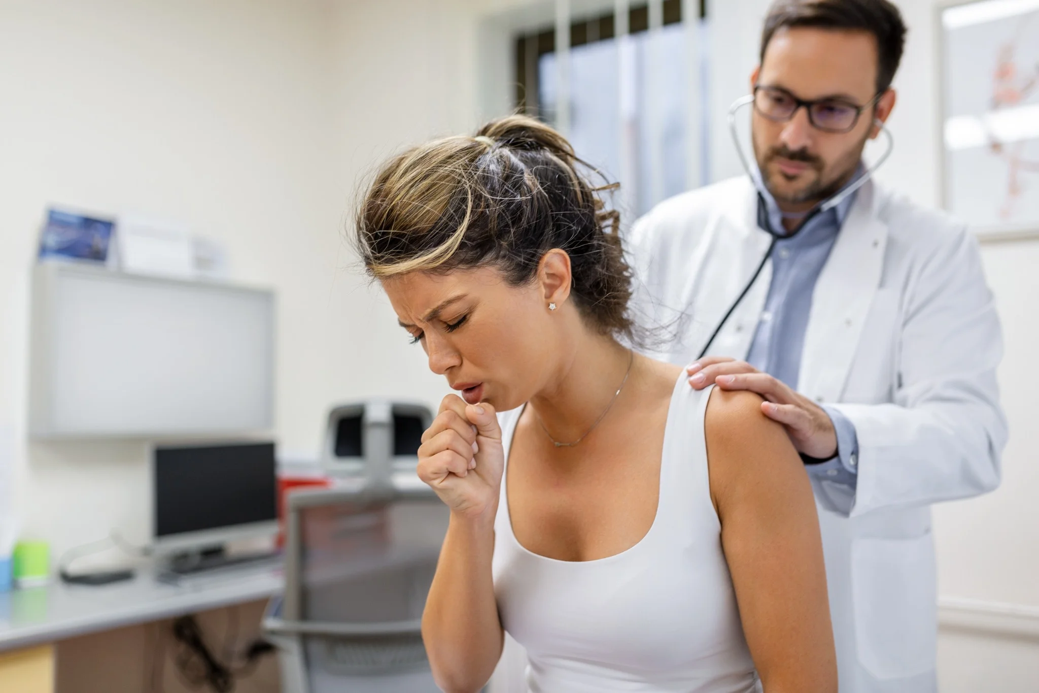 Paciente jovem com tosse, enquanto médico escuta os pulmões com estetoscópio, indicativo de sintomas de pneumonia silenciosa.